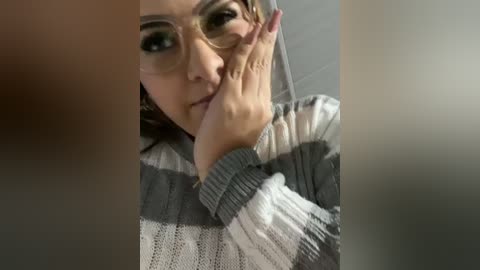 Media: A close-up video of a young woman with fair skin and light brown hair, wearing glasses, a grey and white striped sweater, and holding her hand to her mouth in a thoughtful pose.