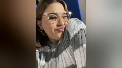 Media: Video of a young woman with light brown skin, wearing clear glasses and a striped sweater, playfully sticking out her tongue. Background is blurred.