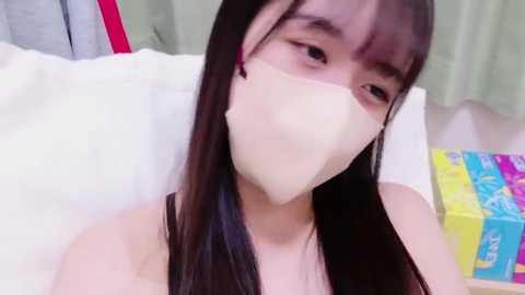 Media: Video of a young Asian woman with long, straight black hair, wearing a beige face mask, lying on a white bed. Background features a colorful box of tissues and light-colored curtains.