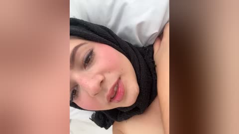 Media: Video of a smiling woman with fair skin, wearing a black headscarf and light makeup, lying on a white pillow in a bedroom.