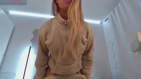 Media: A video of a blonde woman in a beige zip-up hoodie, standing indoors with white walls and a modern, minimalist decor.