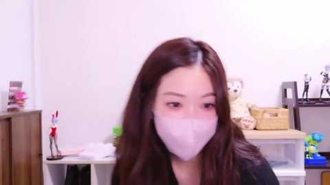 Media: A video of a young East Asian woman with long, straight brown hair and light skin, wearing a white surgical mask, sitting in a cluttered room with white walls, a wooden cabinet, and various toys.