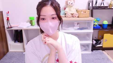 Media: Video of a young Asian woman in a white robe, wearing a face mask, sitting at a desk in a cluttered room with shelves, toys, and office supplies.