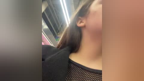 Media: A video of a young woman with light skin, wearing a black mesh top, in a subway car. The background shows fluorescent lighting and a pink jacket.