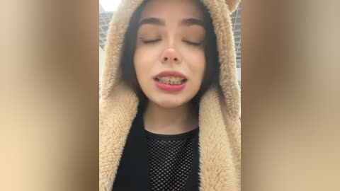 Media: Video of a young woman with fair skin, closed eyes, and braces, wearing a beige, fluffy hooded costume with a black mesh top. Background is blurred.