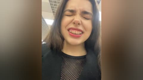 Media: A close-up video of a young woman with straight, dark hair and fair skin, wearing a black mesh top and a black blazer, smiling with braces. She stands in a brightly lit office with visible ceiling tiles.