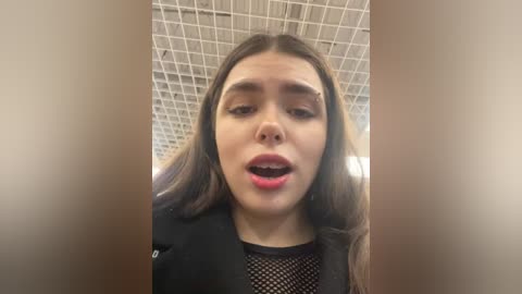 Media: Video of a young woman with long brown hair, wearing a black mesh top, captured from a low angle, with a grid-tiled ceiling background.