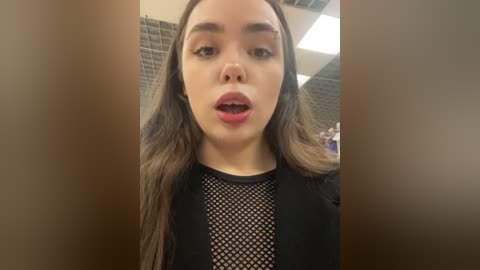 Media: Video of a young, light-skinned woman with long, dark hair, wearing a black fishnet top, standing in a tiled bathroom with fluorescent lighting.