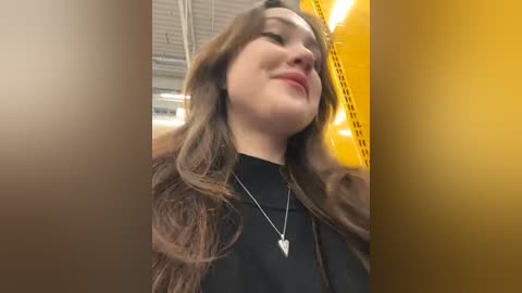 Media: Video of a smiling young woman with long brown hair, wearing a black top and necklace, standing inside a yellow taxi. Background shows a blurred indoor setting with metal ceiling panels.