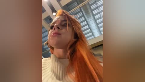 Media: Video of a fair-skinned woman with long, vibrant orange hair, wearing a white, ribbed turtleneck sweater. She has a confident expression, and the background shows a modern indoor setting with large windows and metal blinds.