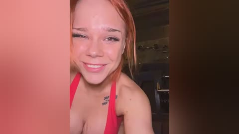 Media: A video of a smiling, light-skinned young woman with red hair, wearing a red halter top, in a dimly-lit kitchen with a blurred background.