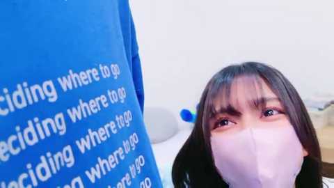 Media: Video of a young Asian woman with straight black hair, wearing a pink mask, standing in front of a blue recycling bin with white text.