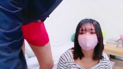 Media: Video of an East Asian woman with shoulder-length black hair wearing a white face mask, kneeling on a bed, looking up at a man in a red briefs and dark pants.