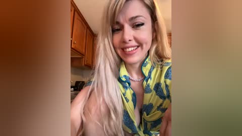 Media: A candid video of a smiling, topless woman with long blonde hair, wearing a yellow shirt with a blue pineapple pattern, in a kitchen with wooden cabinets.