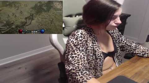 Media: Video of a young woman with brown hair, wearing a leopard print jacket over a black bra, seated at a wooden desk in a modern room with grey carpet.