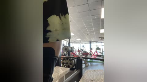 Media: Video of a modern gym with high ceilings, bright lights, and a person wearing a black shirt and shorts working out on a weight machine.