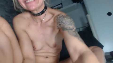 Media: Video of a topless, slender, blonde-haired woman with a black choker, smiling. She has a tattooed arm and a pale skin tone. Background includes a bed with a dark blanket and a white wall.