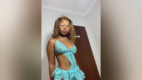 Media: Video of a young Black woman with shoulder-length curly hair, wearing a sheer turquoise lingerie set with cutouts, standing in a minimalist room with a brown door.