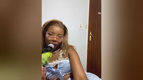 Media: Video of a light-skinned Black woman with braids, wearing glasses, a blue dress, and holding a green plastic bottle, in a dimly lit room with a wooden door.