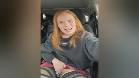 Media: Video of a smiling woman with long, straight red hair, wearing a grey sweatshirt and pink shorts, sitting in a car. She has fair skin, and her left hand is suggestively placed on her crotch.