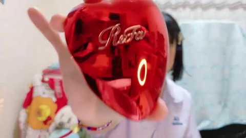 Media: Video of a hand holding a shiny red heart-shaped balloon with a lit-up 'LOVE' design, set against a blurred background of a white curtain and colorful fabric.