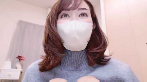 Media: Video of a young woman with shoulder-length auburn hair, wearing a blue turtleneck sweater, white face mask, and clear glasses, gazing upwards. Background features light-colored walls and a grey curtain.