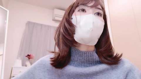 Media: Video of an East Asian woman with shoulder-length brown hair, wearing a white surgical mask, light blue sweater, and beige wall background.