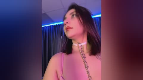 Media: Video of a pale-skinned woman with straight brown hair, wearing a pink spaghetti strap top and a white choker with a silver chain. Background features blue and purple lighting with dark curtains.