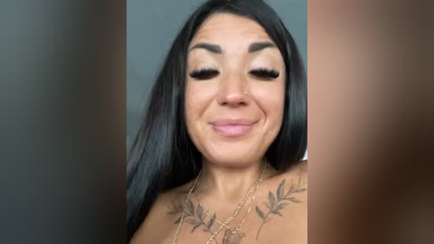 Media: Video of a smiling woman with long black hair, wearing black eyeliner, mascara, and nude lipstick. She has a tattoo of leafy vines on her chest and wears a gold necklace.