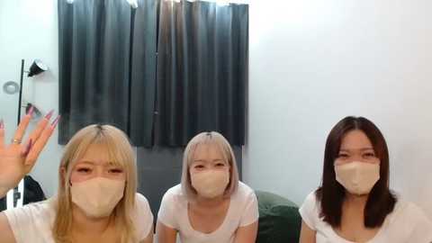 Media: Video of three young women, all wearing white masks, seated in a changing room. One raises her hand in a wave. Background features grey curtains and a green couch.