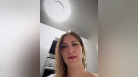 Media: Video of a fair-skinned woman with long, straight, light brown hair, wearing minimal makeup, standing in a modern, white-walled room with a ceiling light fixture.