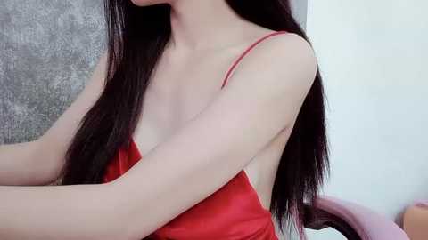 Media: Video of a young woman with long, straight black hair, wearing a red spaghetti-strap dress, sitting indoors. The background features a textured gray wall and a white wall.