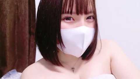 Media: A video of a young Asian woman with a short bob haircut, wearing a white mask, off-shoulder white top, and a delicate necklace. Background shows a wooden wall and blurred white surface.