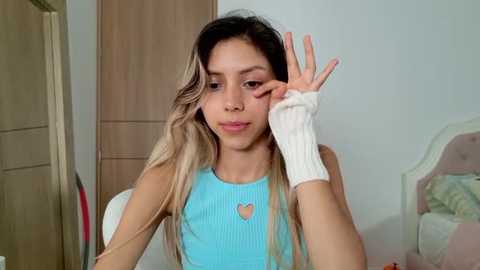 Media: Video of a young Latina woman with long, wavy, blonde hair, wearing a light blue ribbed tank top with a heart pendant. She's seated indoors, making a peace sign with her right hand, while her left hand rests on a white arm cuff.