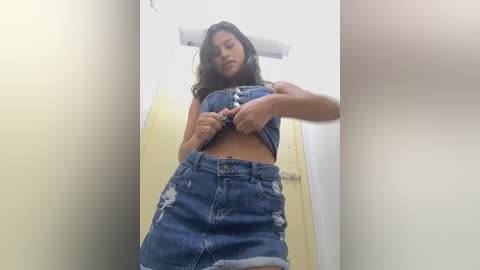 Media: Video of a young woman with long brown hair, wearing a blue, distressed denim crop top and high-waisted shorts, standing in a narrow, brightly lit corridor.