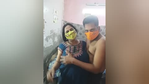 Media: A video shows a shirtless man with a yellow mask, holding a woman in a blue patterned dress with a yellow mask, sitting on a bed in a dimly lit, cluttered room.