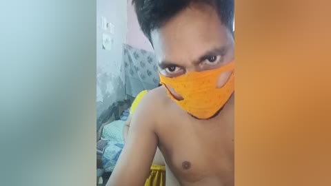 Media: A video shows a shirtless, young South Asian man with medium skin tone, wearing an orange face mask and a yellow cloth around his waist. The background features a cluttered room with a gray couch and a blue-patterned cushion.