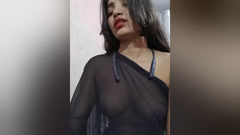 Media: Video of a woman with long dark hair, wearing a sheer black top, showing her breasts and nipples. The background is blurry and out of focus.