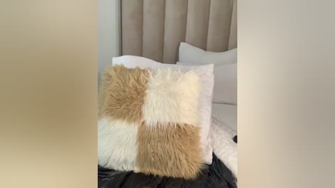 Media: A video of a plush, square, multi-colored faux fur pillow with beige, brown, and white patches on a bed with white pillows and a black blanket. The headboard is beige and padded.