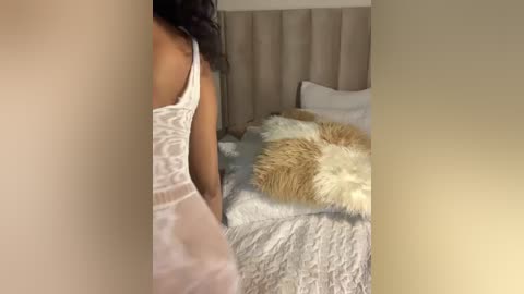 Media: Video of a woman in white lace lingerie standing beside a beige upholstered bed with white and brown faux fur throw.