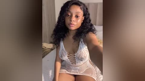 Media: Video of a young Black woman with medium-brown skin, wearing a revealing white lace lingerie dress, sitting on a bed with a beige headboard, in a softly lit bedroom.