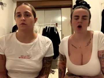 Media: Video of two young women with fair skin, one with light brown hair in a bun, the other with dark hair pulled back, both wearing tight white crop tops, in a bedroom with a closet and clothes hanging.