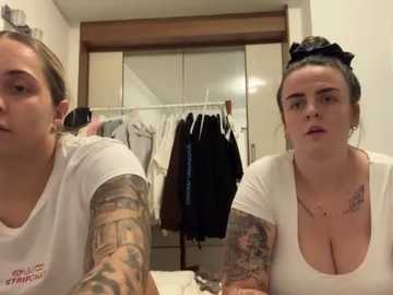 Media: Video of two women with tattoos, wearing white shirts, in a cluttered bedroom with hanging clothes.