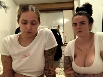 Media: Video of two young women in a bedroom; one with a white crop top, tattoos, and blonde hair, the other with a low-cut top, dark hair, and piercings.