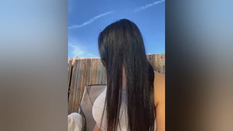 Media: Video of a woman with long, straight black hair, partially covering her face, wearing a white tank top. She is standing outdoors near a wooden fence under a clear blue sky.