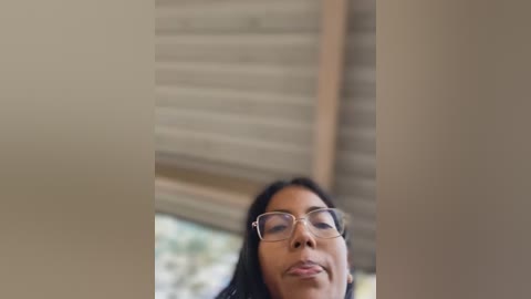 Media: Video of a middle-aged woman with light skin, long black hair, and glasses, smiling with a slight pout. She stands in front of a wooden shuttered window. The background is out of focus.