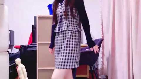 Media: A video of a woman in a black and white checkered dress, standing in a cluttered room with beige shelves, a computer, and a white curtain.