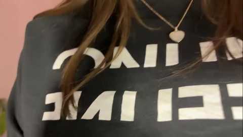 Media: Video of a person wearing a black hoodie with white text that reads \"MADRID\" partially visible. The person has long, dark brown hair and wears a gold necklace with a heart pendant. The background is a blurred, pink wall.