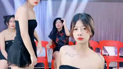 Media: Video of four Asian women in black outfits, one with red makeup, standing against a white backdrop with red chairs, creating a dramatic and modern stage setting.