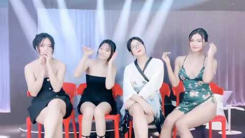 Media: Video of four Asian women with short black hair, seated on red chairs, in a modern, dimly lit room with white curtains, wearing black and green dresses, smiling and gesturing.